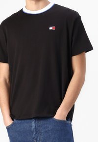 Mies, joka on pukeutunut mustaan Tommy Jeans -t-paitaan, jossa on sininen ja valkoinen kauluskirjailu, sekä sinisiin farkkuihin, kädet taskuissa, valkoista taustaa vasten.