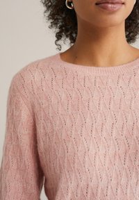 Pulover tricotat de culoare roz deschis, cu un model de valuri, decolteu rotund și textură moale, cu cusături vizibile și o ușoară transparență.