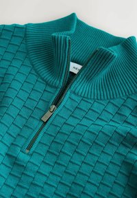 Türkis gestrickter Pullover mit einem Reißverschlusskragen, der ein strukturiertes Quadratmuster und einen gerippten Kragen aufweist. Der Reißverschluss hat eine silberne Farbe.