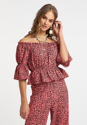 Femeie care poartă un top roz cu umerii goi și pantaloni asortați cu imprimeu negru leopard, accesorizată cu coliere colorate și cercei cu ciucuri.