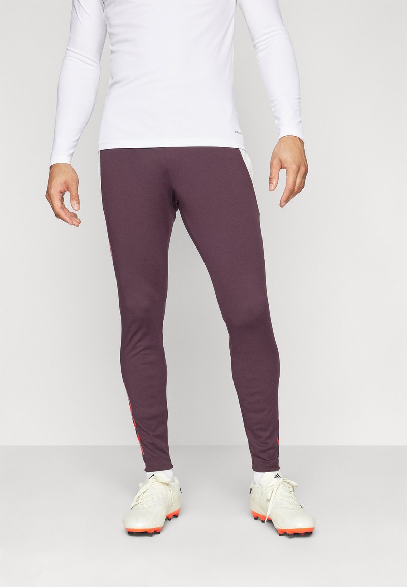Leggings deportivos burdeos hechos de tela elástica, con acentos rojos a los lados y un diseño ajustado. Se llevan con una camiseta de manga larga blanca y botas de fútbol.