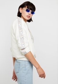 Hvid bomberjakke med broderet blomsterdetalje langs ærmerne, ribbede manchetter og kant. Bæret med lyseblå denim. Stilfulde solbriller.