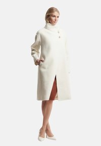 Cappotto bianco in misto lana con colletto alto, caratterizzato da uno spacco frontale, due bottoni e tasche laterali. Il modello indossa un abito a coste sotto.