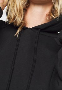 Sudadera negra con cordones y bolsillo frontal. El material parece suave con una textura lisa. Presenta un diseño clásico y un estilo casual.