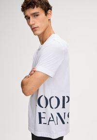 Weißes Baumwoll-T-Shirt mit lockerer Passform, kurzen Ärmeln und auffälligem, navyfarbenem Text "LOOP JEANS" an der Seite. Glatte Textur, keine Muster.