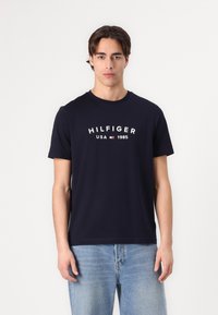 Tamno plavi pamučni majica kratkih rukava s okruglim izrezom i bijelim logotipom "HILFIGER USA 1985" u središtu.