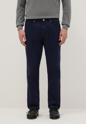 Hombre con pantalones azul marino de corte recto, camiseta gris de manga larga y zapatos negros con cordones, de pie frente a un fondo liso.