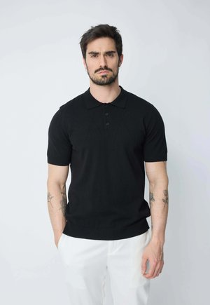Polo noir à manches courtes en tissu tricoté avec un col à trois boutons et des poignets côtelés, associé à un pantalon clair.