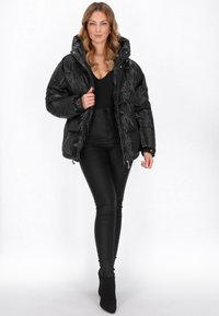 Czarna kurtka puchowa o błyszczącej fakturze, z kapturem i kieszeniami zapinanymi na zamek. Połączona z czarnymi legginsami i botkami za kostkę. Neutralne tło.