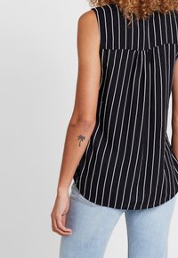 Svart ärmlös topp med vita vertikala ränder, med detalj av bakfåll och rundad nederkant. Bärs med ljusblå jeans.