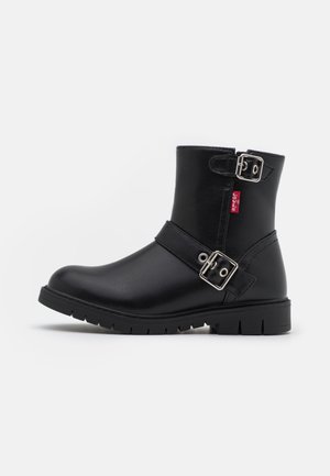 Bottine en cuir noir avec deux boucles argentées et une étiquette rouge sur le côté, avec une semelle robuste et une finition lisse.