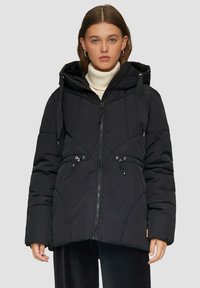 Zwarte pufferjack met rits aan de voorkant, gequiltde textuur, verstelbare trekkoorden en een capuchon. Gemaakt over een coltrui.