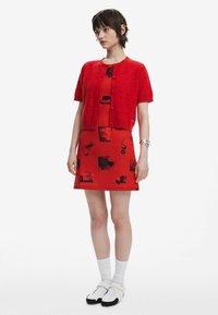 Röd stickad kofta över en röd klänning med svarta grafiska mönster. Vita strumpor och slip-on skor fulländar outfiten. Släta texturer genomgående.