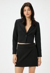 Blazer noir cropped avec une fermeture à bouton et de grands revers crantés, associé à une mini-jupe noire ajustée. Texture lisse, design épuré.