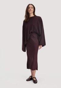 Vrouw in een losse donkerpaarse trui, bijpassende midi rok en donkerpaarse Mary Jane schoenen, staand tegen een effen achtergrond.