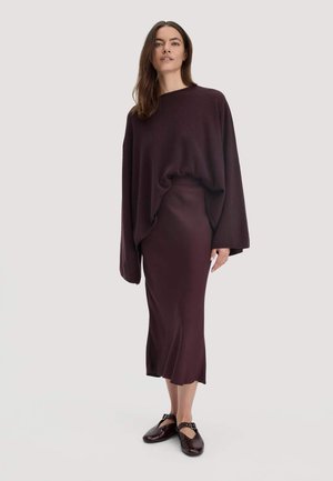 Femme portant un pull ample violet foncé, une jupe midi assortie et des chaussures Mary Jane violettes foncées, se tenant devant un fond uni.