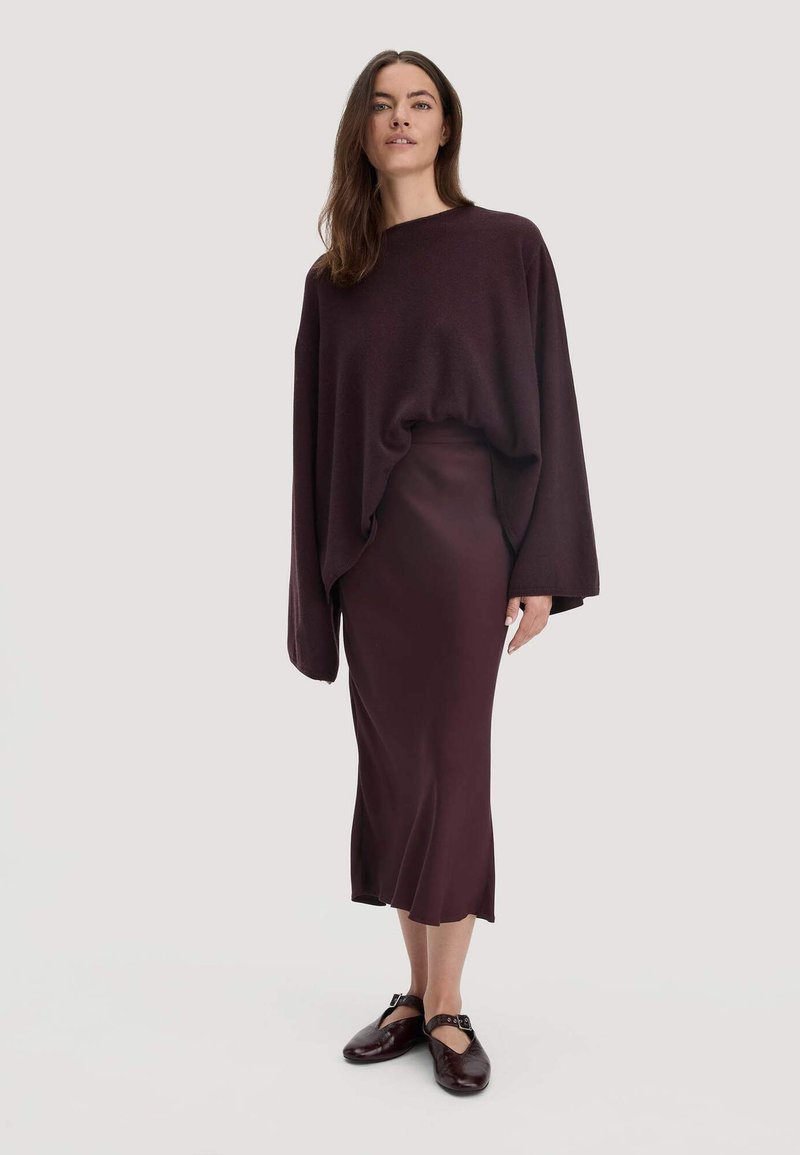 Vrouw in een losse donkerpaarse trui, bijpassende midi rok en donkerpaarse Mary Jane schoenen, staand tegen een effen achtergrond.