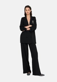 Blazer doppiopetto nero con scollatura a V profonda, abbinato a pantaloni neri larghi e ballerine grigie. Spicca la spilla argento sul blazer.