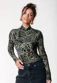 Frau mit braunen Haaren in einem lockeren Dutt, die goldene Ohringe, ein grünes Tiermuster-langärmliges Oberteil und schwarze hochgeschnittene Jeans trägt.