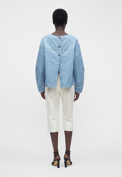 Personne debout de dos portant une veste en jean bleu clair boutonnée, un pantalon blanc court et des sandales noires à talons sur fond uni.