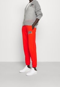 Grå cardigan med knappar och logodetalj, tillsammans med ljusorangea joggers med broderad logotyp, och vita sneakers.