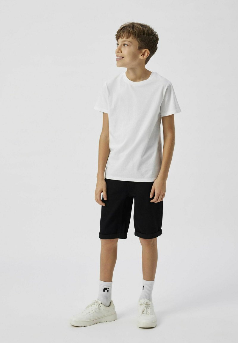 T-shirt en coton blanc, short en denim noir et baskets blanches avec une semelle texturée. Le short est retroussé à l'ourlet, ajusté mais pas serré.