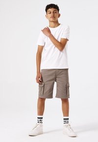 T-shirt blanc, short cargo gris avec plusieurs poches, baskets montantes blanches et chaussettes à motifs noir et blanc. Fond uni.