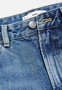 Gros plan de la taille d'un jean en denim bleu avec un bouton en métal et une étiquette de marque MNG visible partiellement rentrée à l'intérieur.