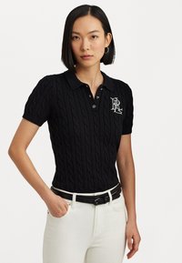 Polo noir en maille avec un motif en relief, manches courtes et col. Présente un logo blanc sur la poitrine gauche. Porté avec un pantalon blanc.