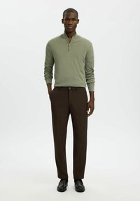 Homme debout, mains dans les poches, portant un pull à demi-fermeture éclair vert olive, un pantalon marron foncé et des chaussures habillées noires sur fond blanc.