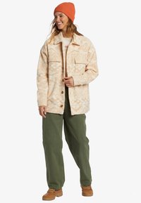 Crème geklede fleece jack met twee borstzakken, knoopsluiting en een oranje gebreide muts. Olijfgroene broek en bruine veterschoenen.