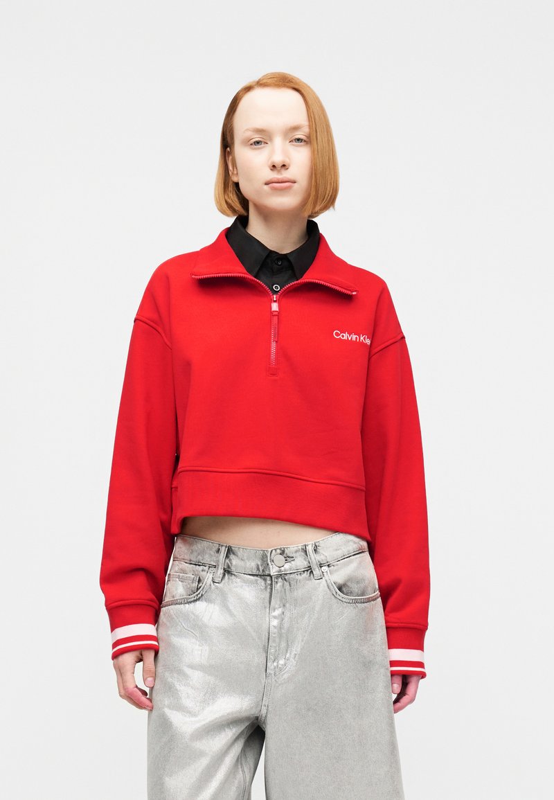 Νεαρός με κόκκινο καρέ κούρεμα, φορώντας κόκκινο κοντό φούτερ με φερμουάρ Calvin Klein και γυαλιστερό ασημένιο ψηλόμεσο παντελόνι.