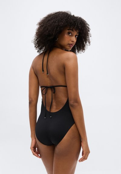 Γυναίκα με σγουρά μαλλιά που φοράει μαύρο ολόσωμο μαγιό με ανοιχτή πλάτη και δεμένες τιράντες, κοιτάζοντας πάνω από τον ώμο της σε απλό φόντο.