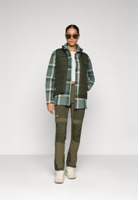 Grüner gepolsterter Weste über einem blau/grünen karierten Hemd, kombiniert mit olivgrünen Cargo-Hosen. Enthält Nähdetails und vordere Taschen, getragen mit weißen Schuhen.
