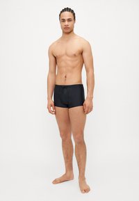Shorts de bain noirs avec un devant plat, une taille ajustable par cordon et une longueur de jambe courte, fabriqués en tissu lisse et extensible, présentés sur un modèle debout.