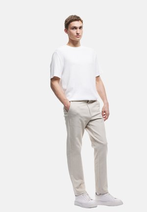 Witte geribbelde t-shirt met korte mouwen, beige taps toelopende broeken en witte sneakers. Model staat met één hand in de zak, neutrale achtergrond.