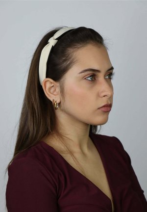 Kremowa opaska na głowę z węzłowym detalem, noszona przez osobę o długich brązowych włosach. Widoczne złote kolczyki-koła oraz głęboko bordowy top.