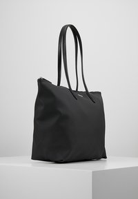 Sac fourre-tout noir en matériau texturé, doté de deux longues poignées et d'un dessus zippé. Design simple avec peu de quincaillerie et d'accents.
