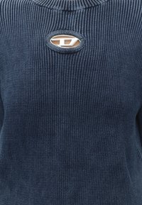 Pull côtelé bleu marine avec une petite découpe ovale sous l'encolure révélant la peau, surmontée d'une boucle décorative argentée.