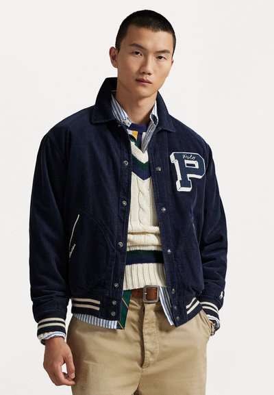 Young man wearing a navy corduroy varsity jacket with a large "P" patch, layered over a cream knit sweater and blue striped shirt, beige pants.