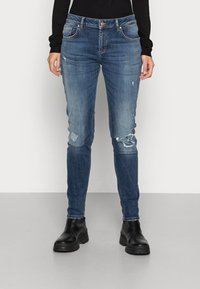 Blå denim skinny jeans med slitna lappar. Har en midje i mellanhöjd, fem fickor och bruna knappar som accent. Bärs med svarta ankelboots.