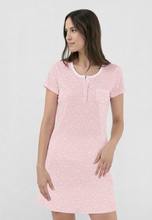 Mujer con un vestido rosa de manga corta con pequeños patrones florales blancos y un bolsillo delantero, mirando hacia su izquierda contra un fondo claro.