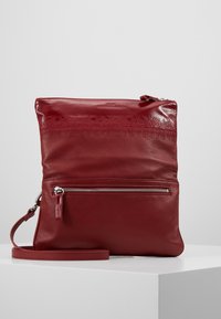 Červená kožená crossbody taška s texturovaným povrchem, horním zipem a přední kapsou se zipem. Má plochý popruh pro nošení.