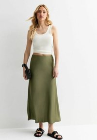 Falda maxi fluida en verde oliva, combinada con una camiseta ajustada blanca. La modelo lleva sandalias planas negras y sostiene un bolso texturizado negro.