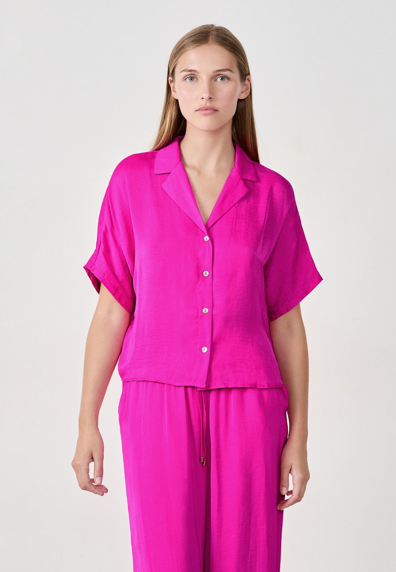 Blusa de manga corta y cuello en fucsia vibrante, hecha de un tejido suave con botones en el frente. Corte holgado con puños ligeramente doblados.