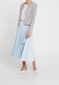 Veste tricotée texturée et multicolore, ouverte à l'avant, assortie à une jupe plissée bleu clair. Des escarpins blancs à bout pointu complètent le look.