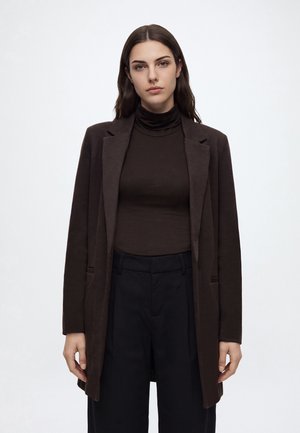 Mujer con cabello largo y oscuro, que lleva una chaqueta marrón oscuro, un cuello alto marrón y pantalones negros, de pie frente a un fondo claro y liso.
