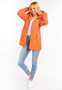 Orange vattentät jacka med huva, framfickor, tryckknappar och justerbar nederkant. Kombinerad med ljusblå jeans och vita sneakers.