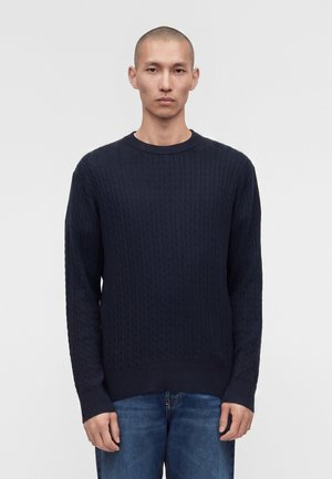 Άνδρας με πολύ κοντό ξύρισμα στο κεφάλι, φοράει σκούνο μπλε πλεκτό πουλόβερ με κόμπους και μπλε τζιν, στέκεται μπροστά σε απλό φωτεινό φόντο.