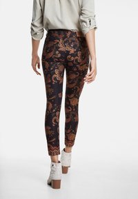 Leggings ajustados con un fondo oscuro que presentan intrincados patrones florales en rojo, dorado y naranja. El modelo lleva botas blancas hasta el tobillo.